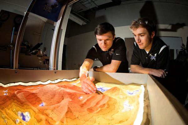 Two individuals in a laboratory setting are observing and interacting with an illuminated topographic sand table. One person reaches out to shape the sand, creating a terrain-like structure with vibrant colors representing elevation and water features. Both wear dark shirts with embroidered logos, focusing intently on the display.